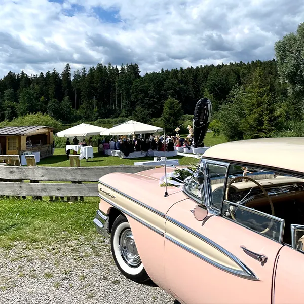 rosafarbenes altes Auto vor Grünanlage mit Hochzeitsgesellschaft und hellen Sonnenschirmen